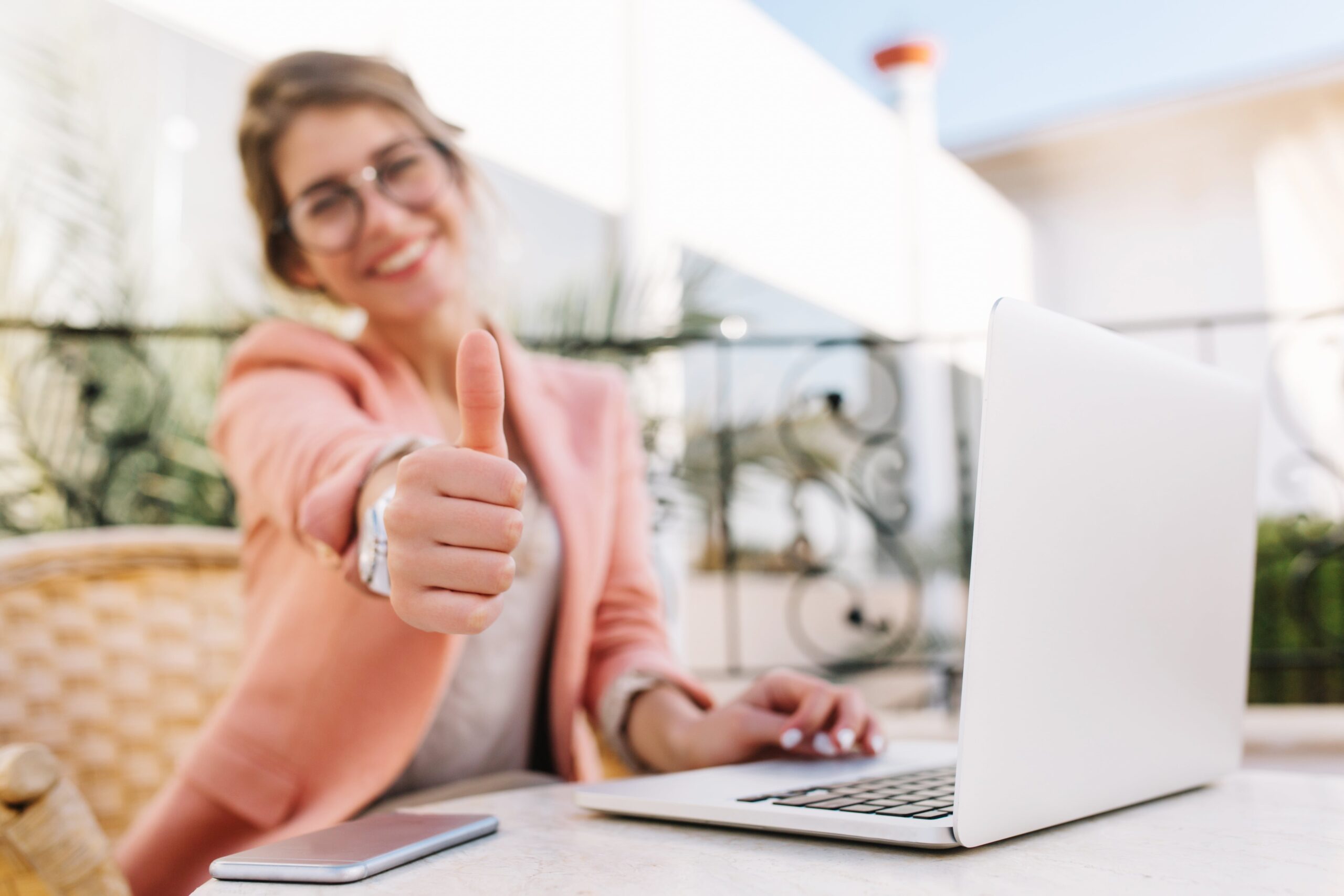 cute-young-woman-student-business-lady-showing-thumbs-up-well-done-sitting-outdoor-cafe-terrace-with-laptop-wearing-pink-smart-clothes-min-scaled
